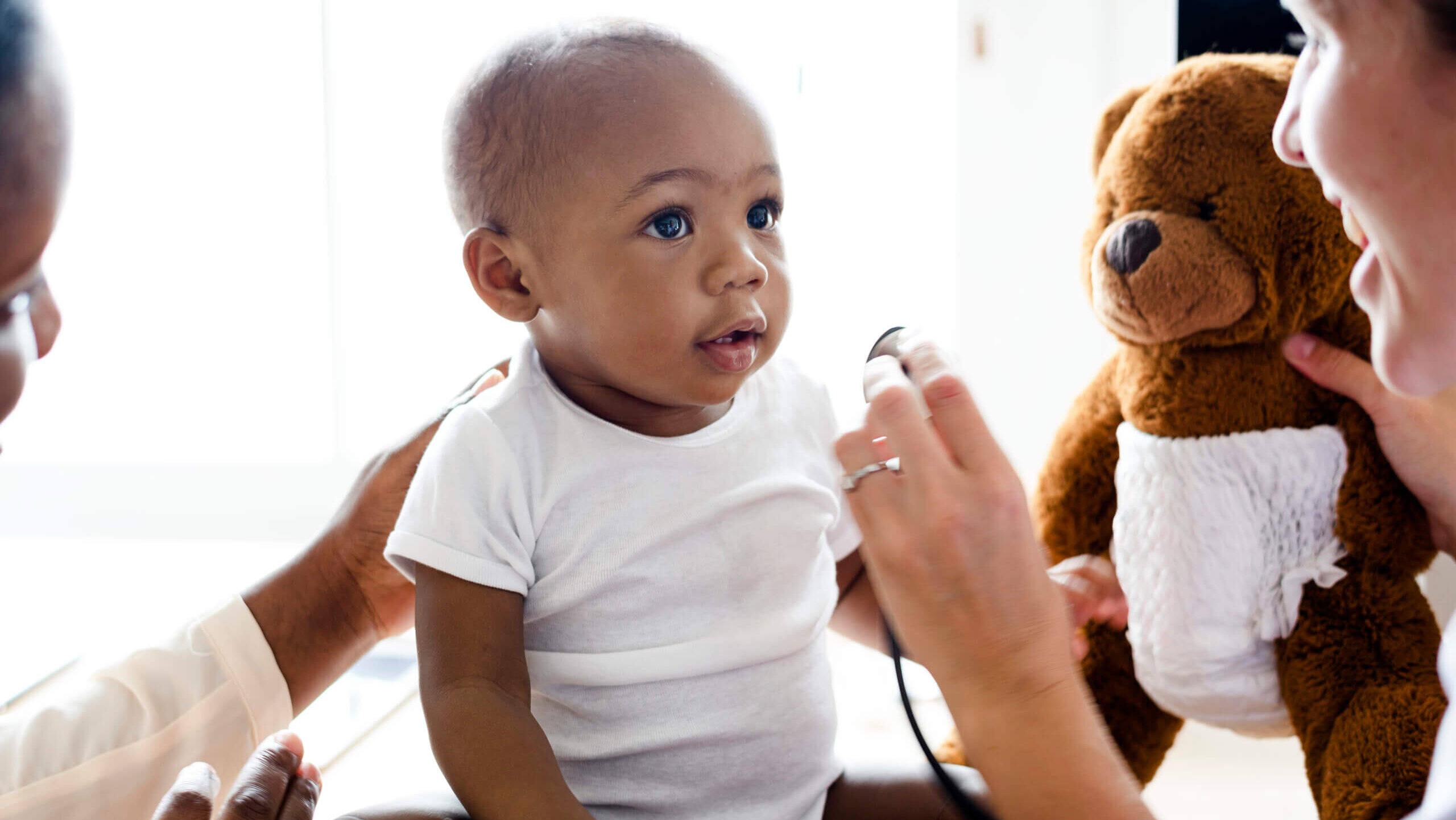Toddler in onesie with doctor and teddy bear.