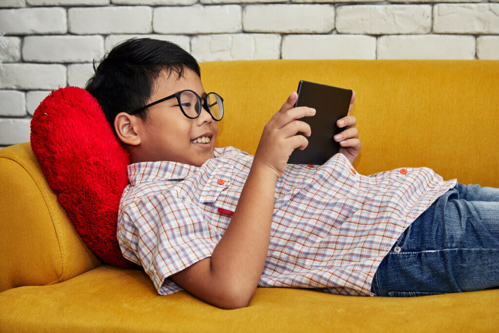 Asian boy smiling playing with tablet wihle laying on yellow sofa
